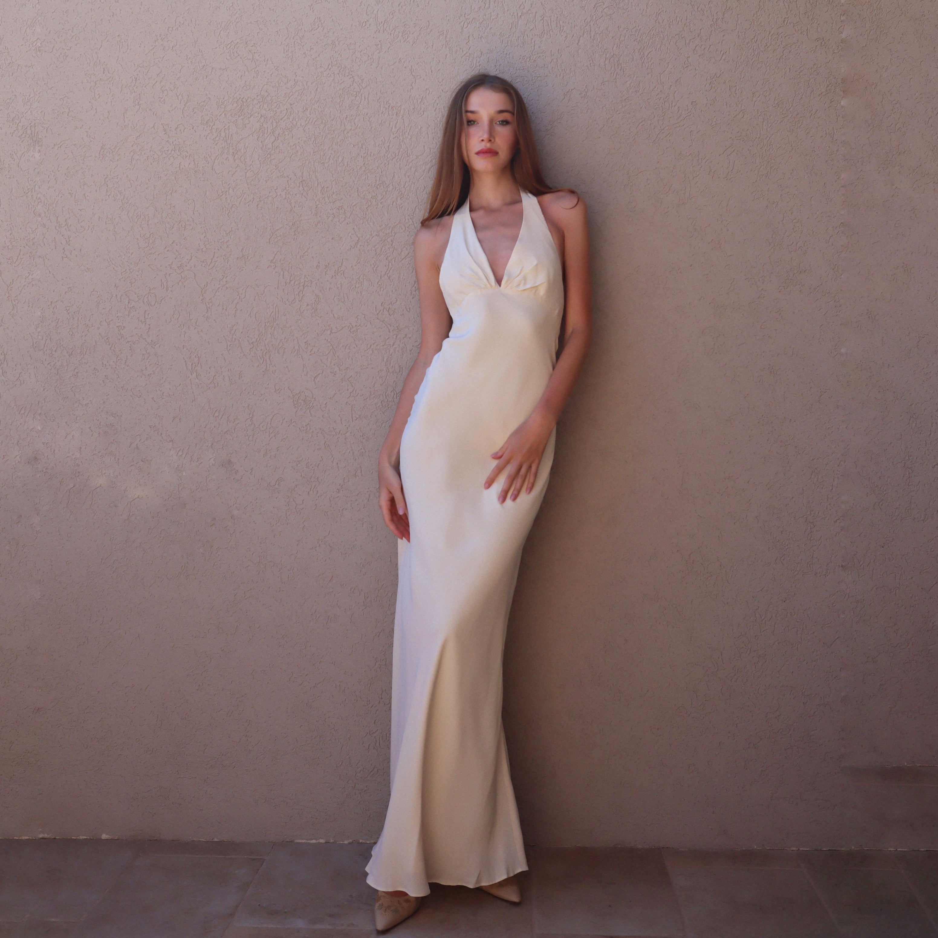 Woman in a white halter neck gown standing against a beige wall.